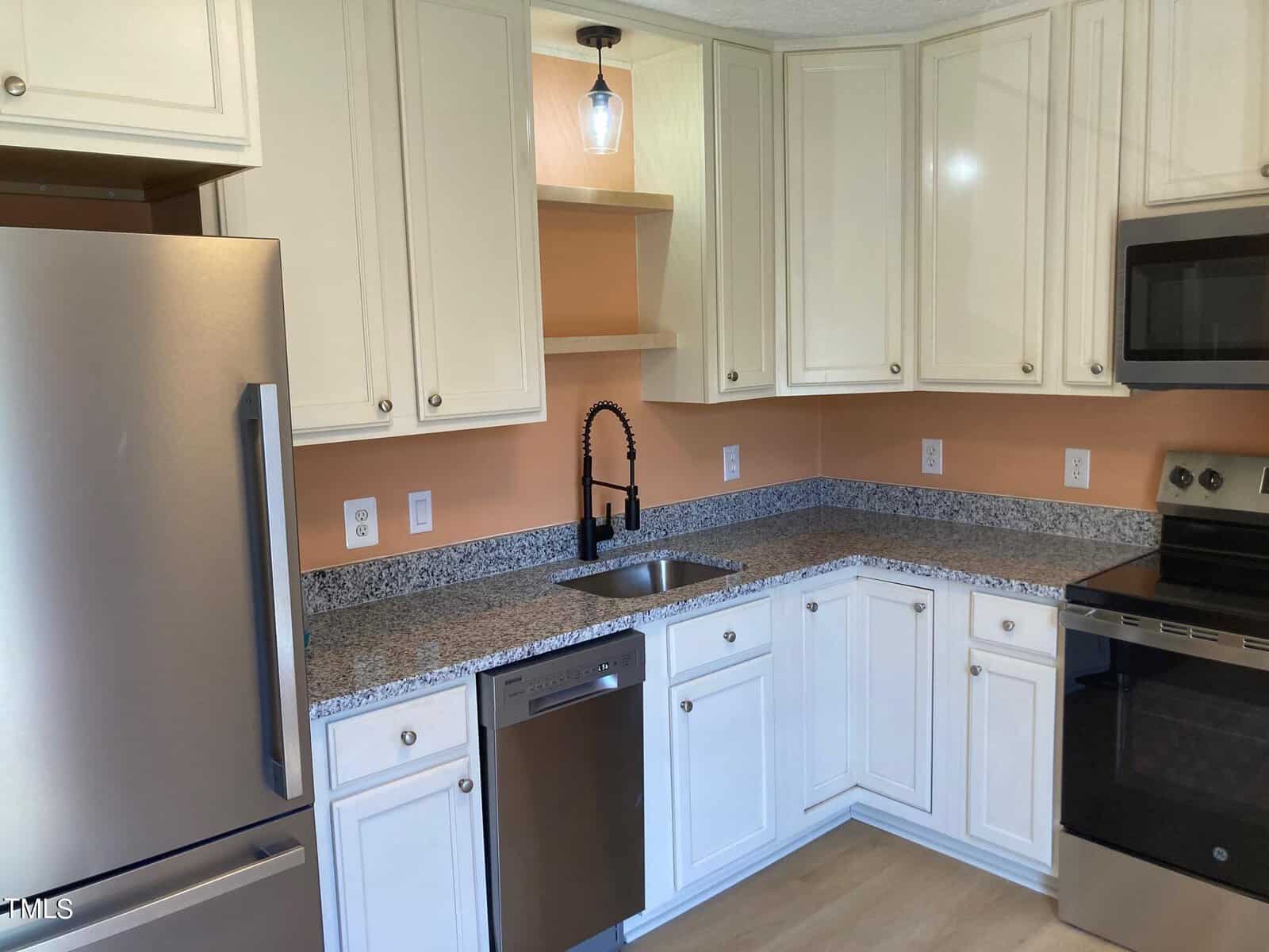 A modern kitchen with white cabinets, stainless steel appliances, and a granite countertop. There's a black faucet over a single-basin sink, and the walls are painted a warm peach color. A small pendant light hangs above the counter.