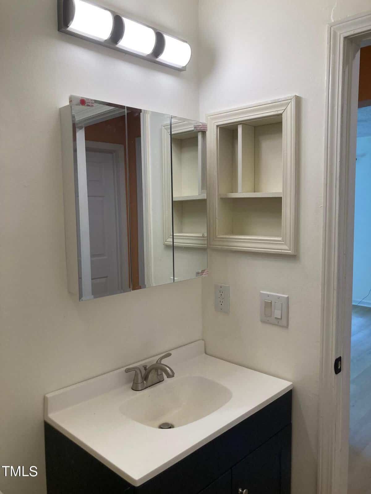 A bathroom features a white sink with a silver faucet on a black cabinet. A large mirrored cabinet is mounted above. The walls are white, with an empty wall shelf and a doorway leading to another room visible in the background.