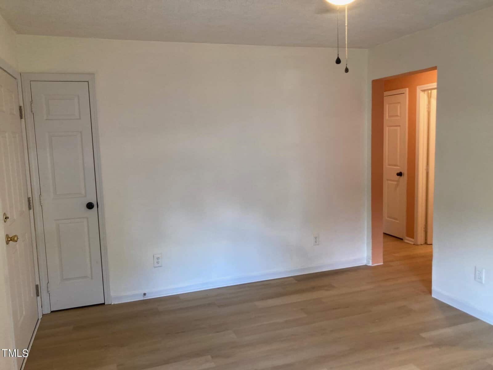An empty room with light wood flooring and white walls. A ceiling fan with a light hangs from the ceiling. A doorway leads to another room with a closed door, and two closed doors are visible on the left wall.