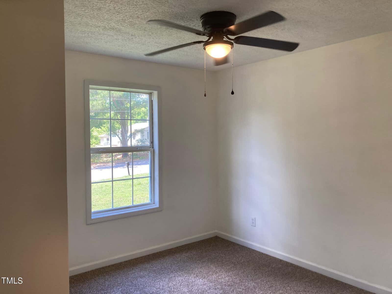 A small, empty room with white walls and a single window showcasing a grassy outdoor area. The room has a ceiling fan with a light and a carpeted floor.
