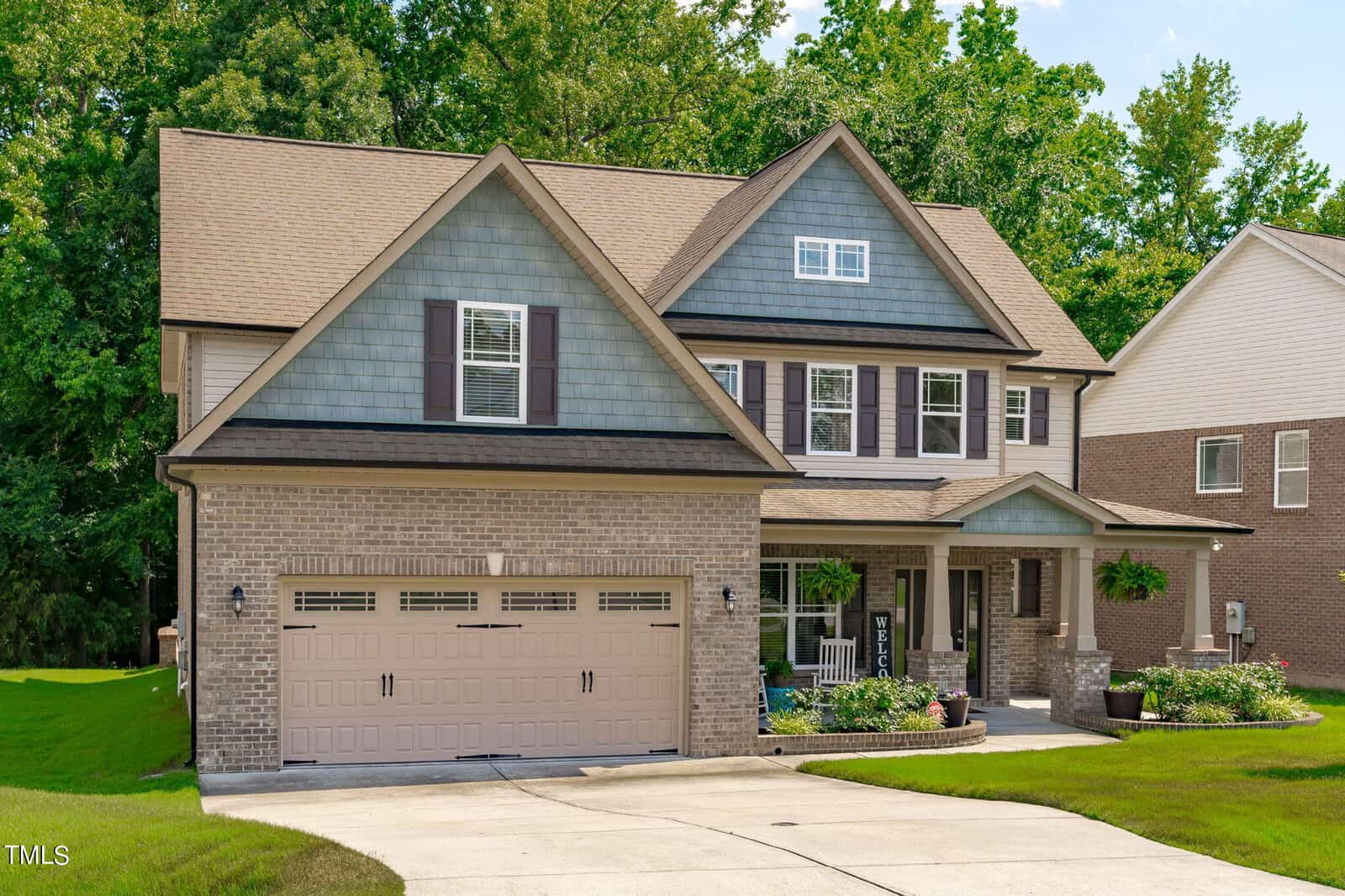 A two-story suburban house with a brick and blue siding facade, featuring a double garage, a small front porch with chairs, and a well-maintained lawn. The background includes dense green trees and a neighboring brick house.