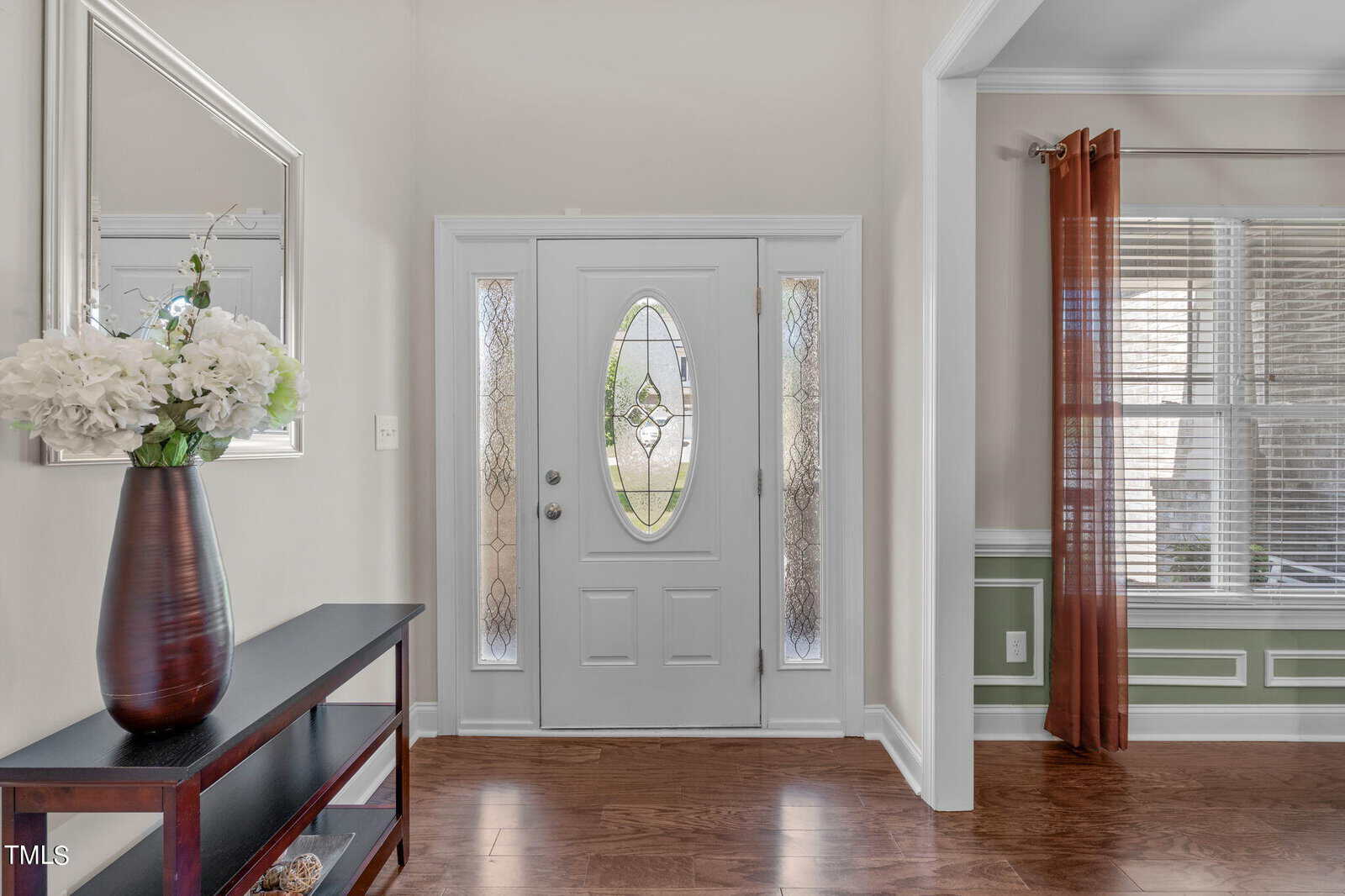 A bright, elegant hallway with a white front door featuring decorative glass panels. A wooden console table with a vase of white flowers sits on the left, and a room with a window and orange curtains is partially visible on the right.