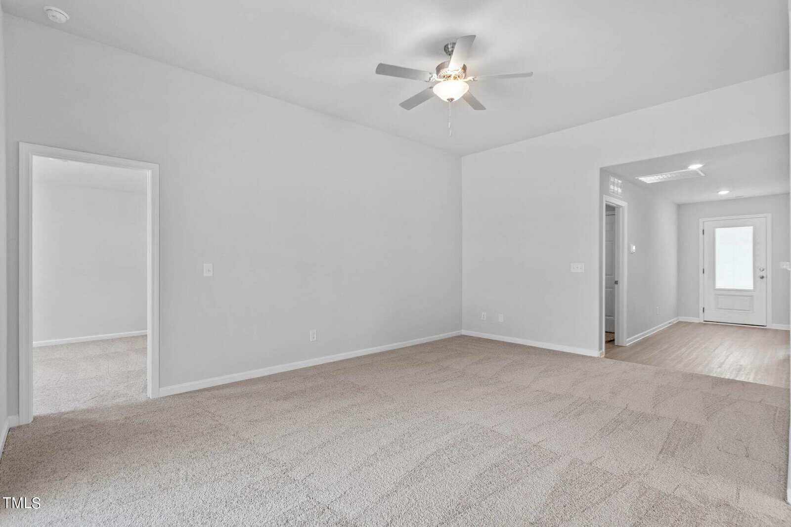 A bright, empty room with beige carpet and white walls. A ceiling fan is centered on the ceiling. Two open doorways lead to adjacent rooms, and a hallway with wooden flooring leads to a white door at the back. Natural light filters in.