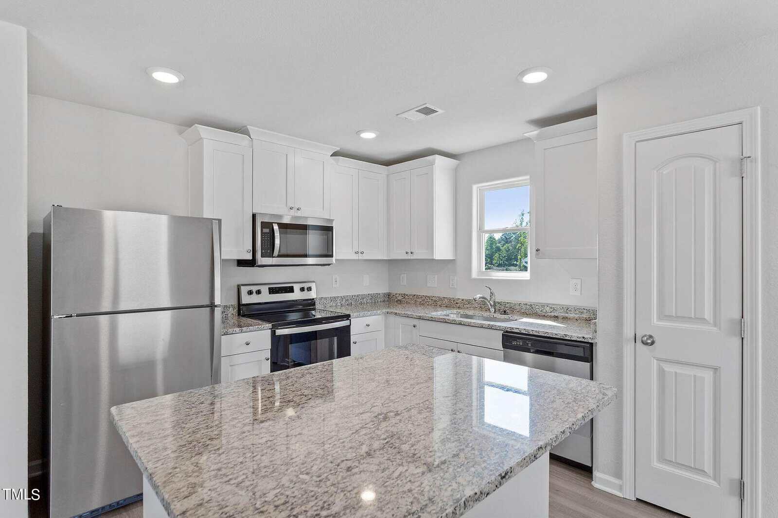 A modern kitchen featuring stainless steel appliances, white cabinets, and a granite countertop island. The space has a window above the sink, providing natural light, and a door to the right.
