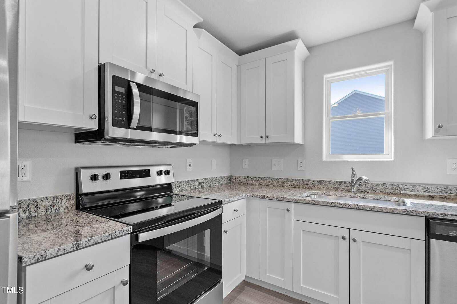 Modern kitchen with white cabinets, granite countertops, and stainless steel appliances, including a stove and microwave. A single window provides natural light, and a sink is installed under the window. Light wood flooring completes the space.