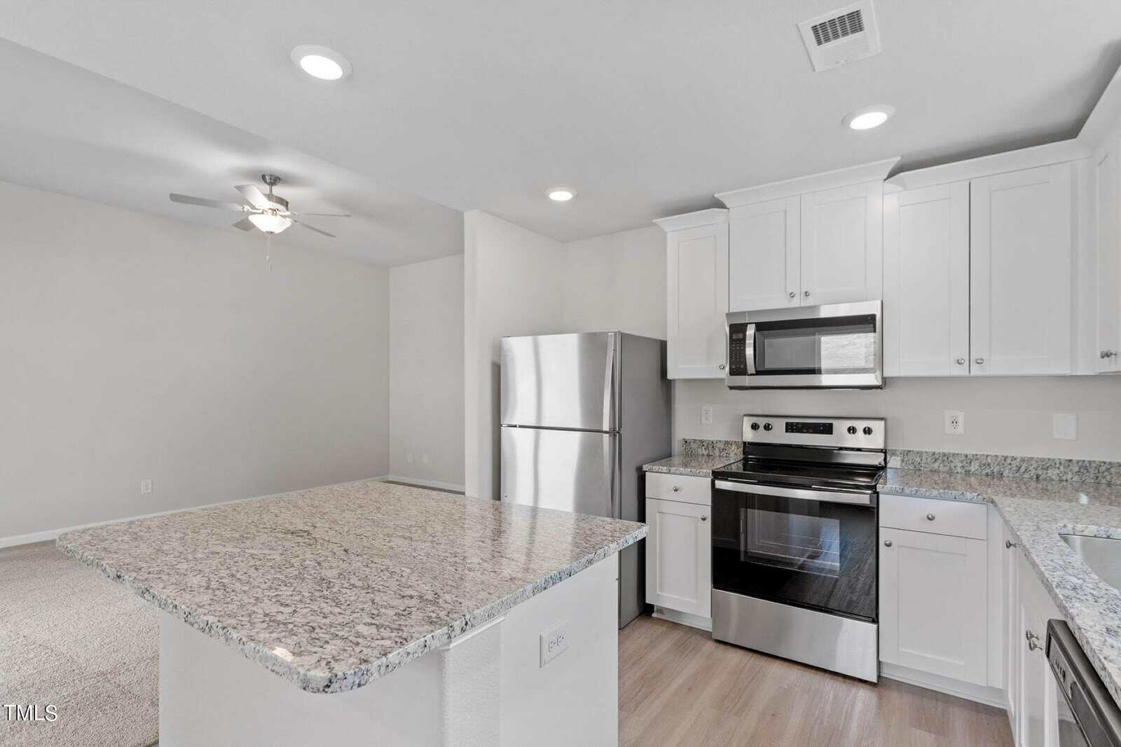 Modern kitchen with white cabinets, granite countertops, and stainless steel appliances, including a fridge, oven, and microwave. An island counter is in the foreground, and a ceiling fan is in the adjacent room. Walls are painted light gray.