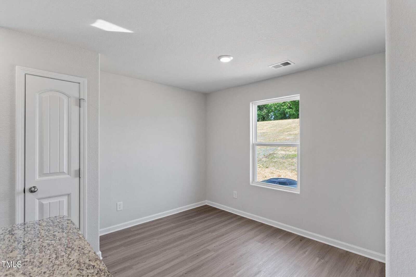 A small, unfurnished room with light gray walls and a wood floor. A single window on the right lets in natural light, showing part of a grassy area outside. A white door is on the left with a doorknob. A small section of granite countertop is visible.