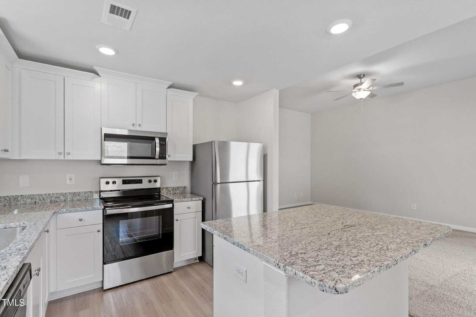 Modern kitchen with white cabinets, stainless steel appliances, and granite countertops. An island with a bar surface is in the foreground, and a ceiling fan is visible in the adjacent open space.