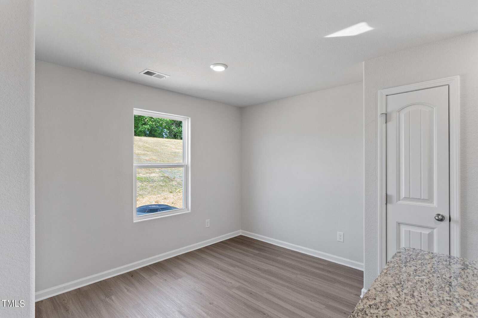 A small, empty room with white walls and a wood floor. A single window lets in natural light, and an interior door is closed. A granite counter is partially visible in the foreground, suggesting a kitchen area nearby.