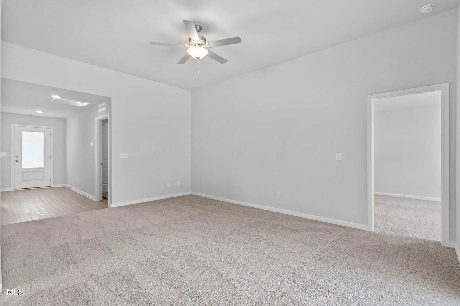 Bright, empty room with light gray walls and carpeted floor. Ceiling fan with light fixture. Doorway leads to a hallway with a view of another room and a glass back door. Minimalist and clean interior design.