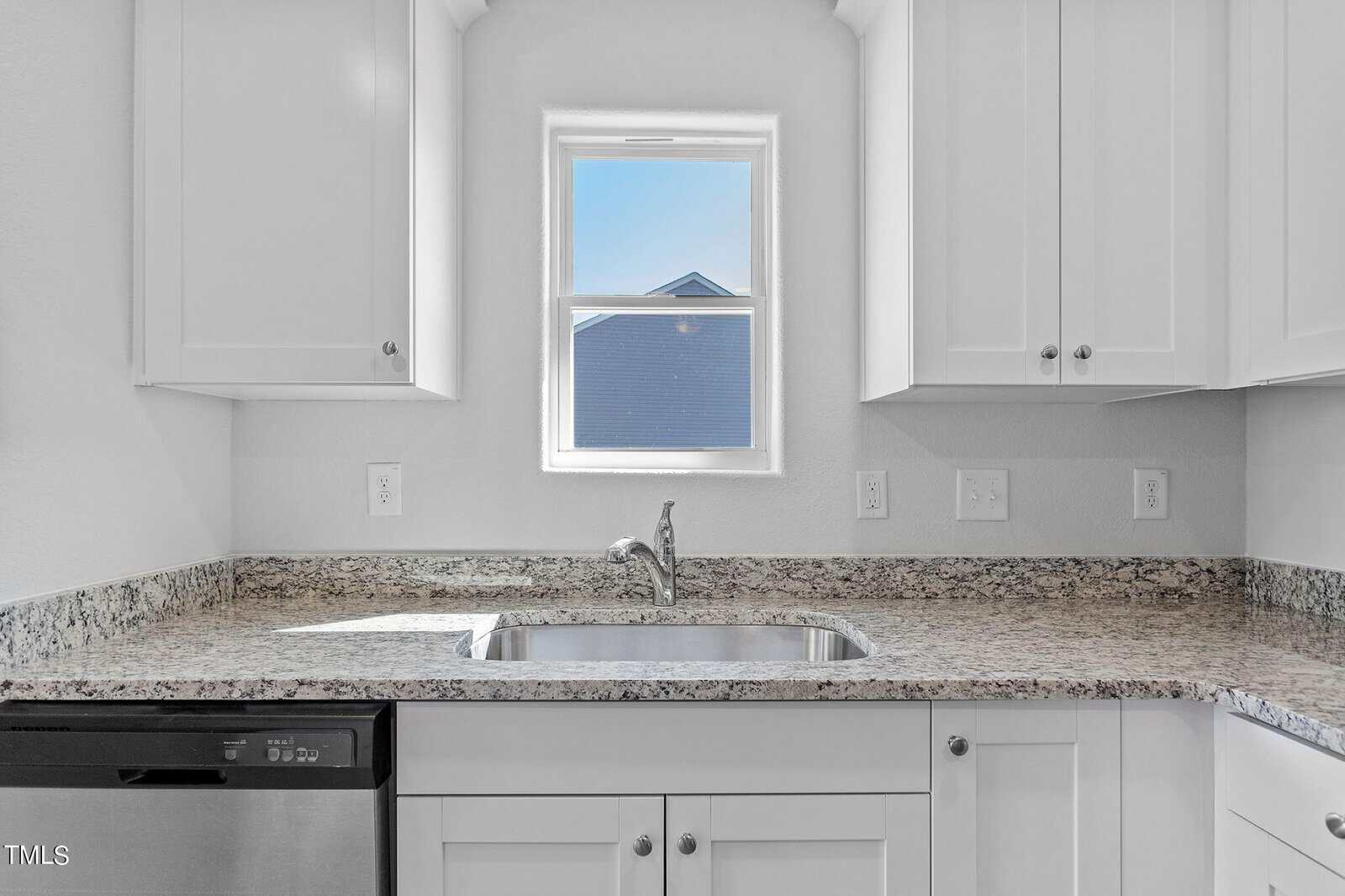 A modern kitchen sink set against a light granite countertop. Surrounding white cabinets frame a small window with a view of a blue house. Stainless steel faucet and dishwasher on the side enhance the sleek, clean design.