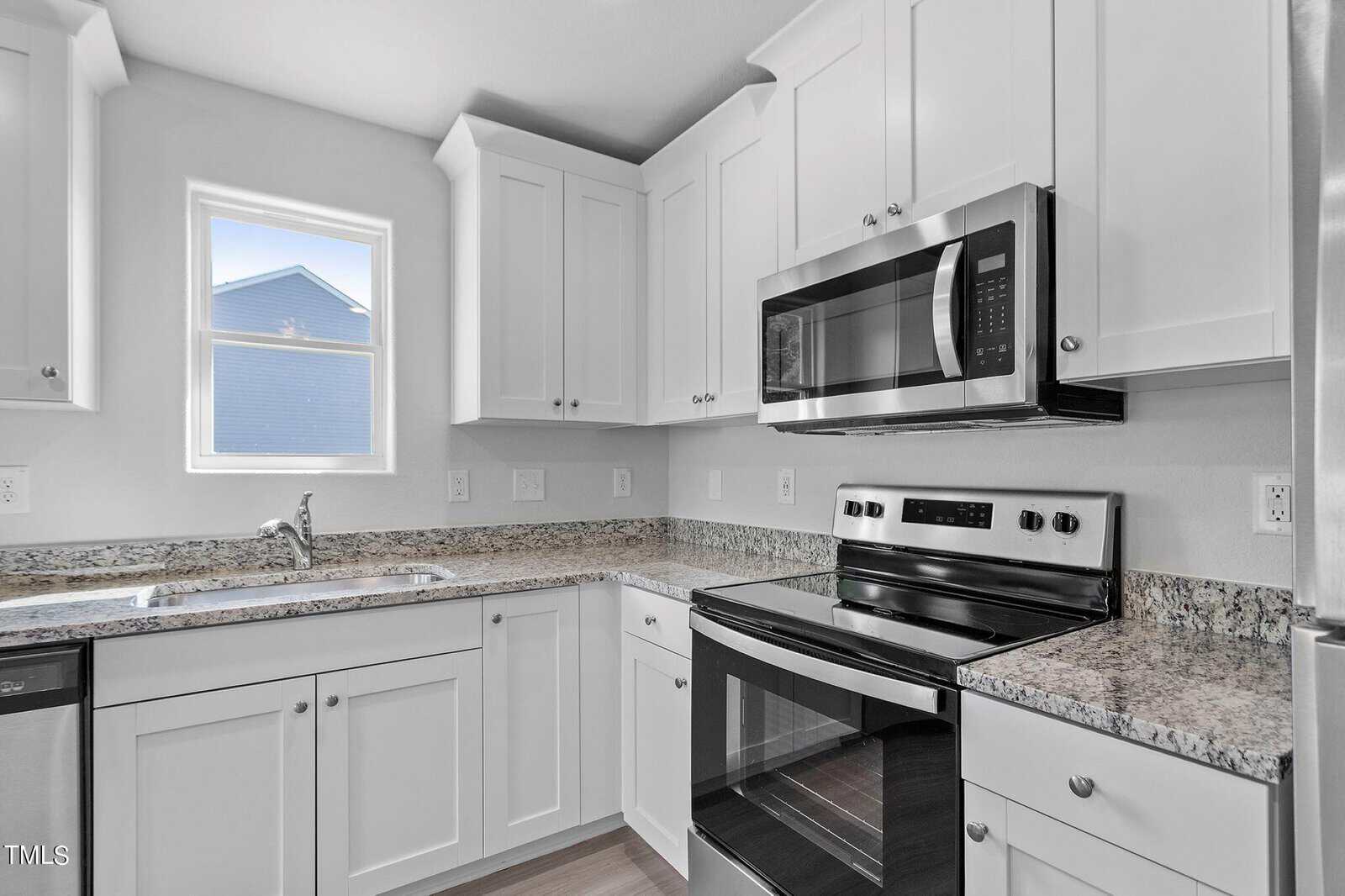 Modern kitchen with white cabinets, granite countertops, and stainless steel appliances including a microwave and oven. A dishwasher is next to the sink under a window. Natural light brightens the space.