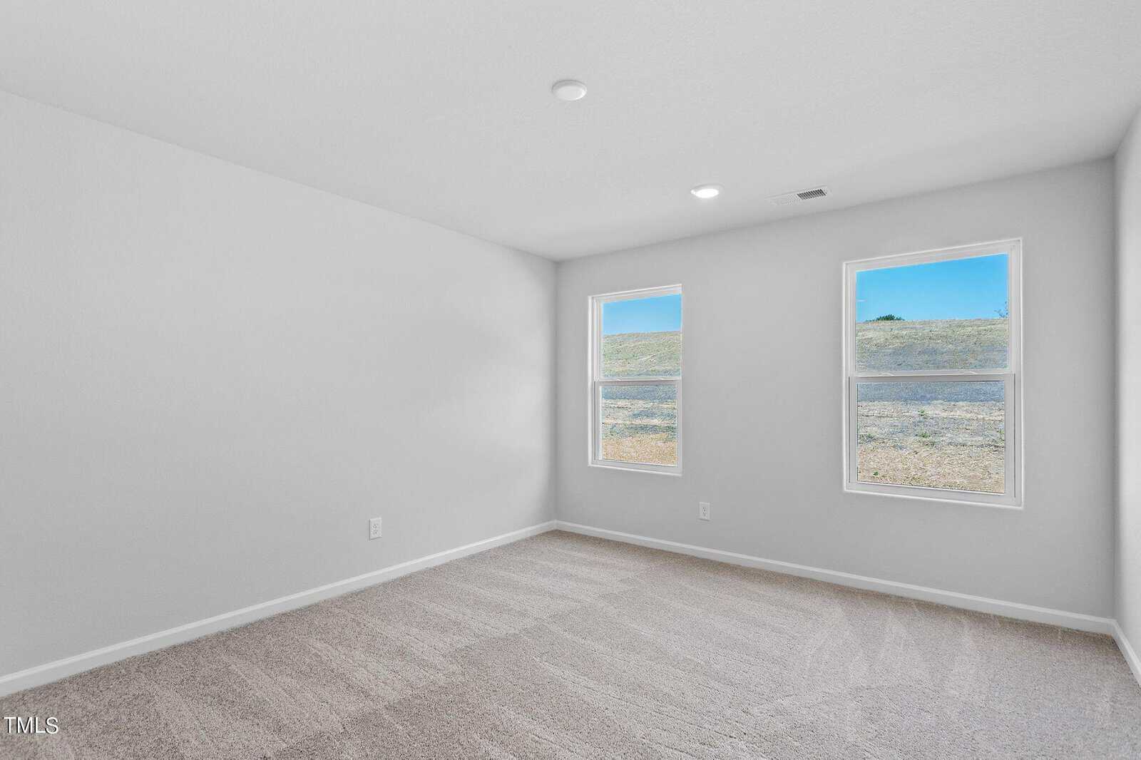 An empty room with light gray walls and carpeted flooring. Two windows on one wall allow natural light, showing an outdoor grassy area and blue sky. The ceiling has recessed lighting.