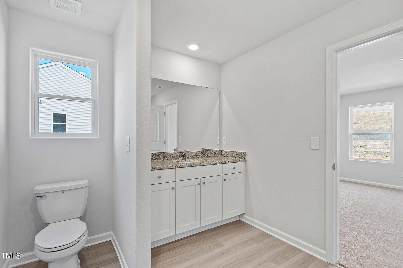 A modern, minimalist bathroom featuring light wood flooring, a white toilet, and a wide mirror above a white vanity with a granite countertop. A window allows natural light, and there's an open doorway leading to a carpeted bedroom.