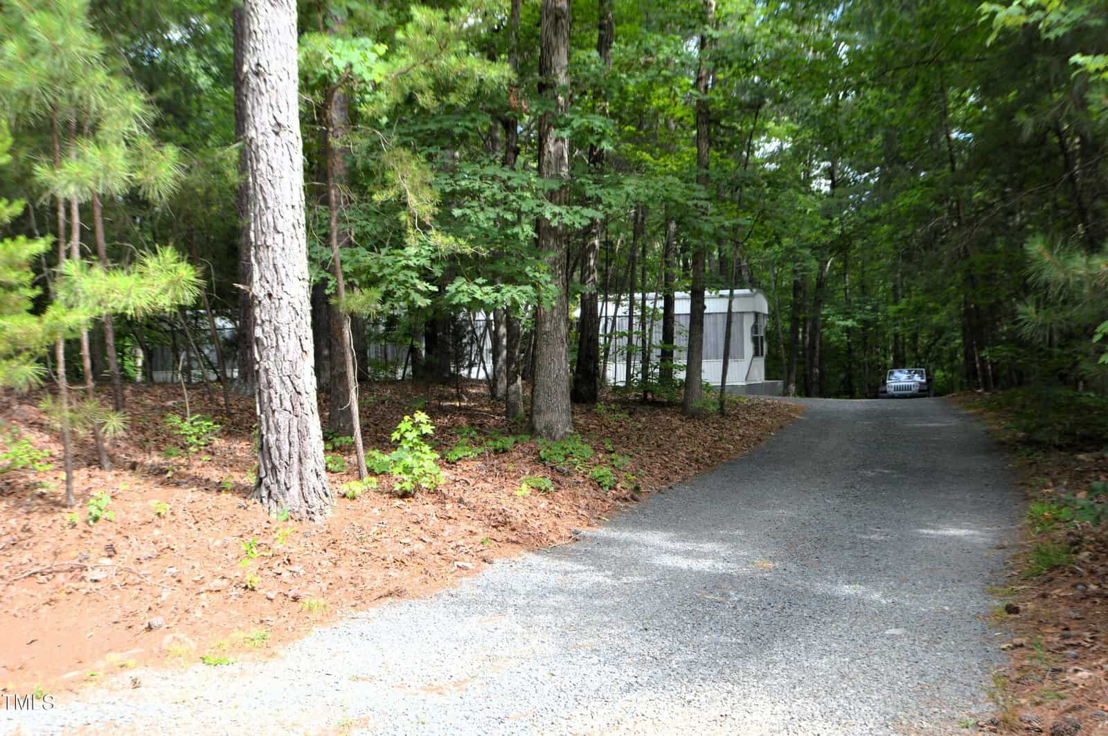A gravel driveway leading through a wooded area with tall trees, towards a white mobile home. A car is parked near the home, surrounded by dense greenery.