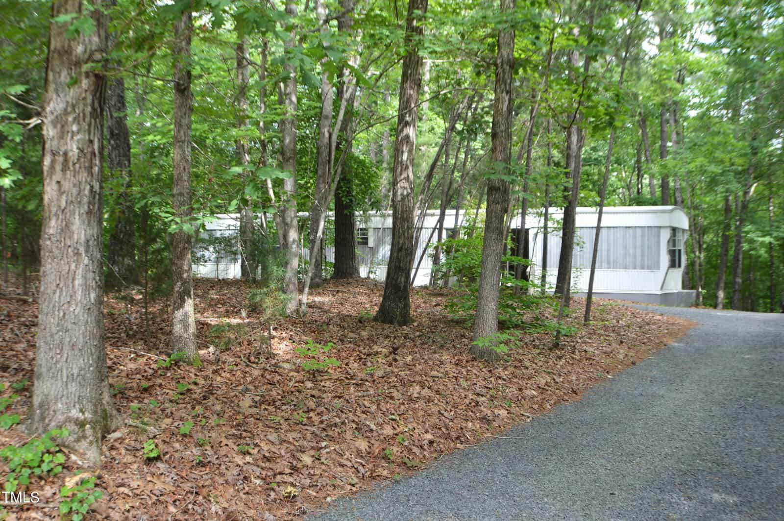 A white mobile home is partially visible through dense green forest trees. A gravel path curves towards the structure, surrounded by a carpet of brown leaves. The scene suggests a quiet, secluded woodland setting.