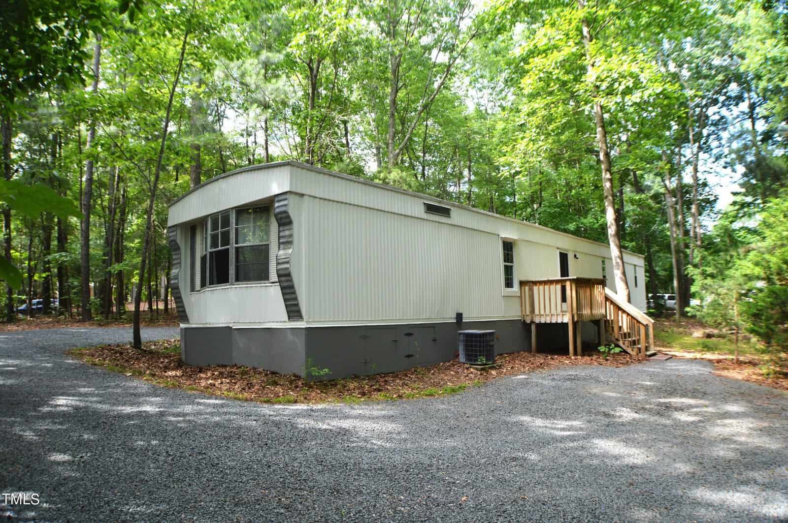A white mobile home with a small wooden porch is surrounded by lush green trees. The home is set on a gravel driveway and has an air conditioning unit on the side. The setting is peaceful and wooded.