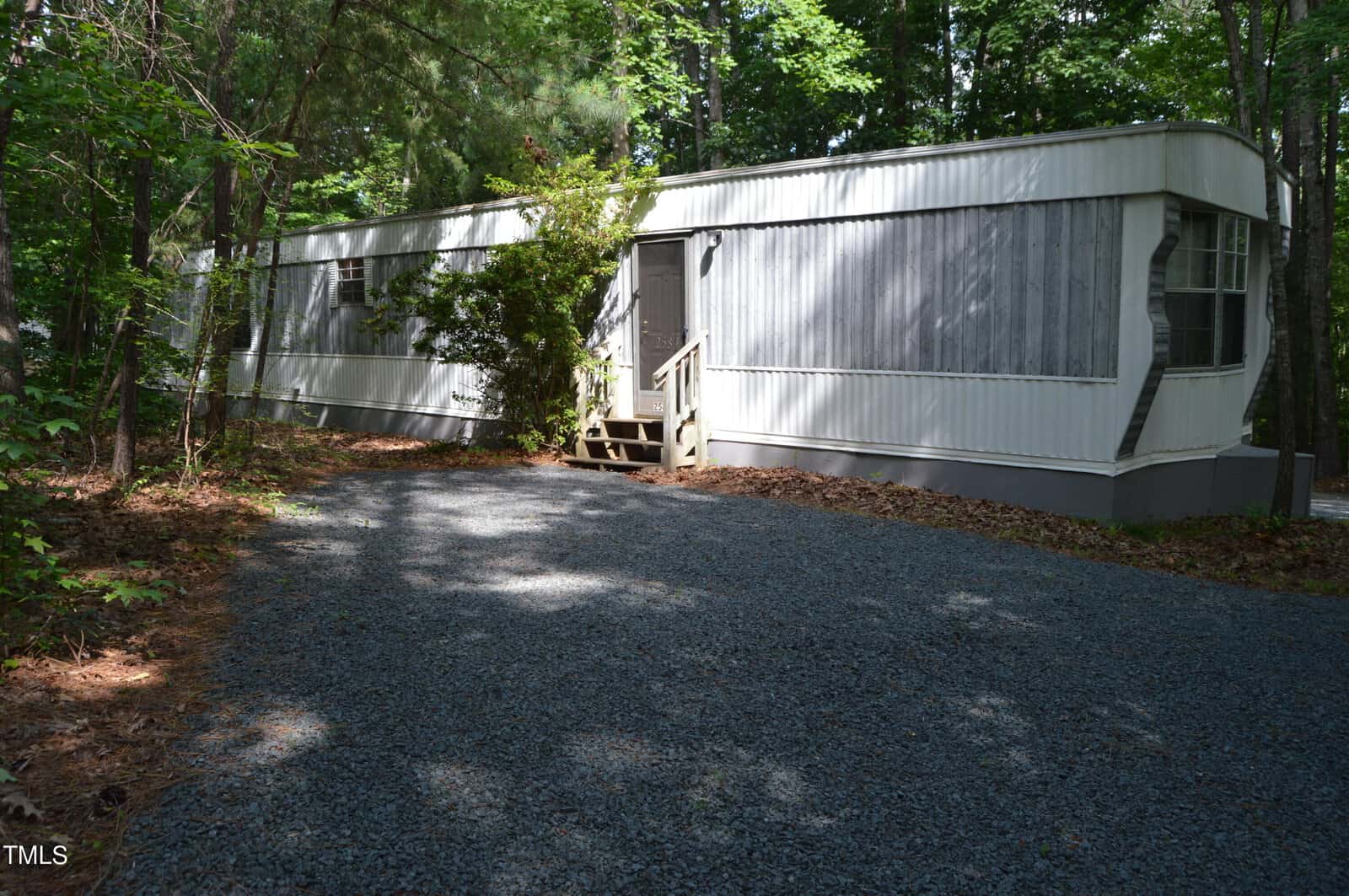 A white mobile home is nestled in a wooded area, surrounded by tall trees. It sits on a gravel driveway, with sunlight filtering through the leaves. A small set of wooden steps leads to the main entrance.