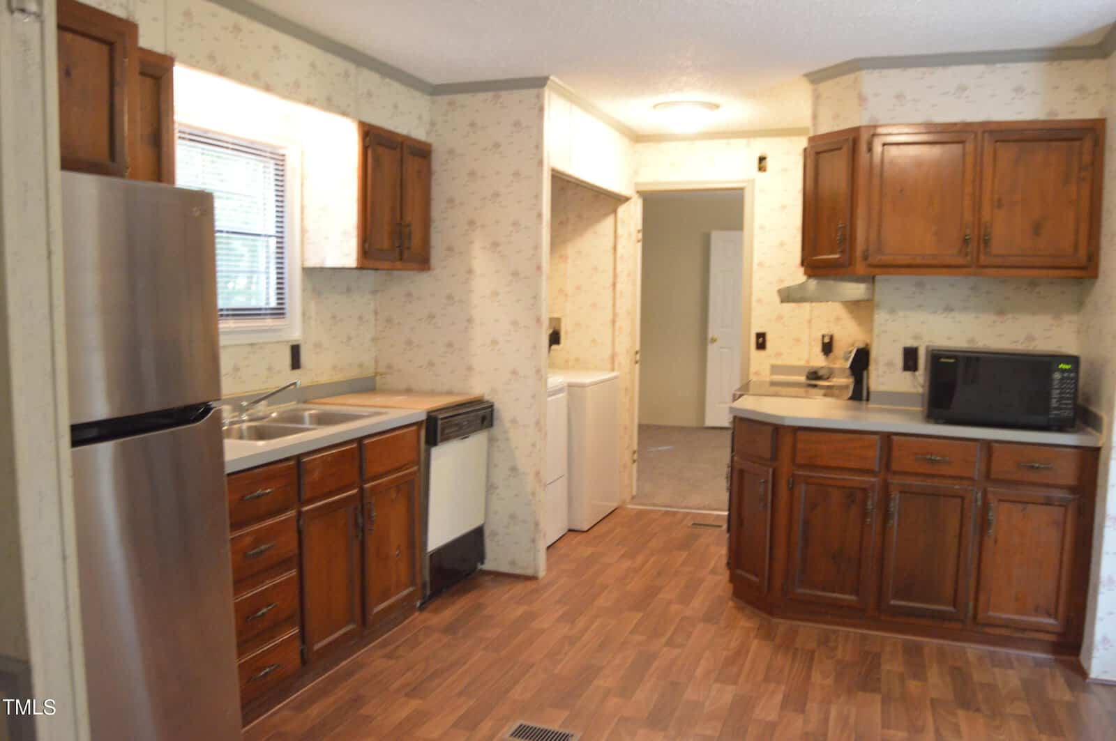 A cozy kitchen with wooden cabinets, stainless steel fridge, sink, and dishwasher. A microwave is on the counter. The floor has wood-patterned vinyl. A small window lets in natural light. Laundry area is visible in the background.