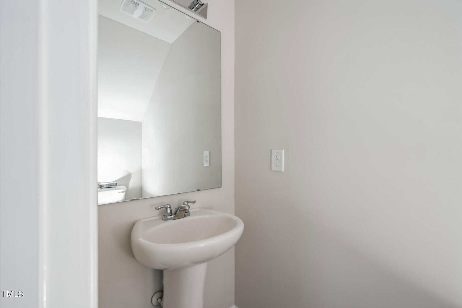 A small bathroom corner featuring a white pedestal sink with a chrome faucet, a rectangular mirror above the sink, and a light switch on the adjacent wall. The walls are painted a light gray color.