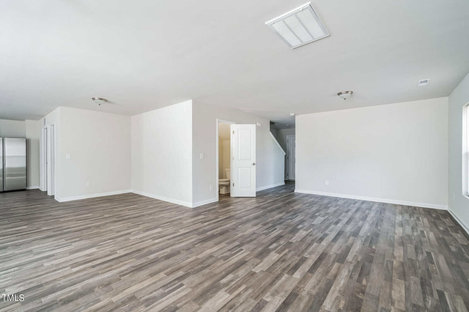 A spacious, empty room with light-colored walls and dark vinyl plank flooring. The room features a small alcove leading to a white door, next to a closed storage area. In the background, part of a stainless steel refrigerator is visible in another open space.