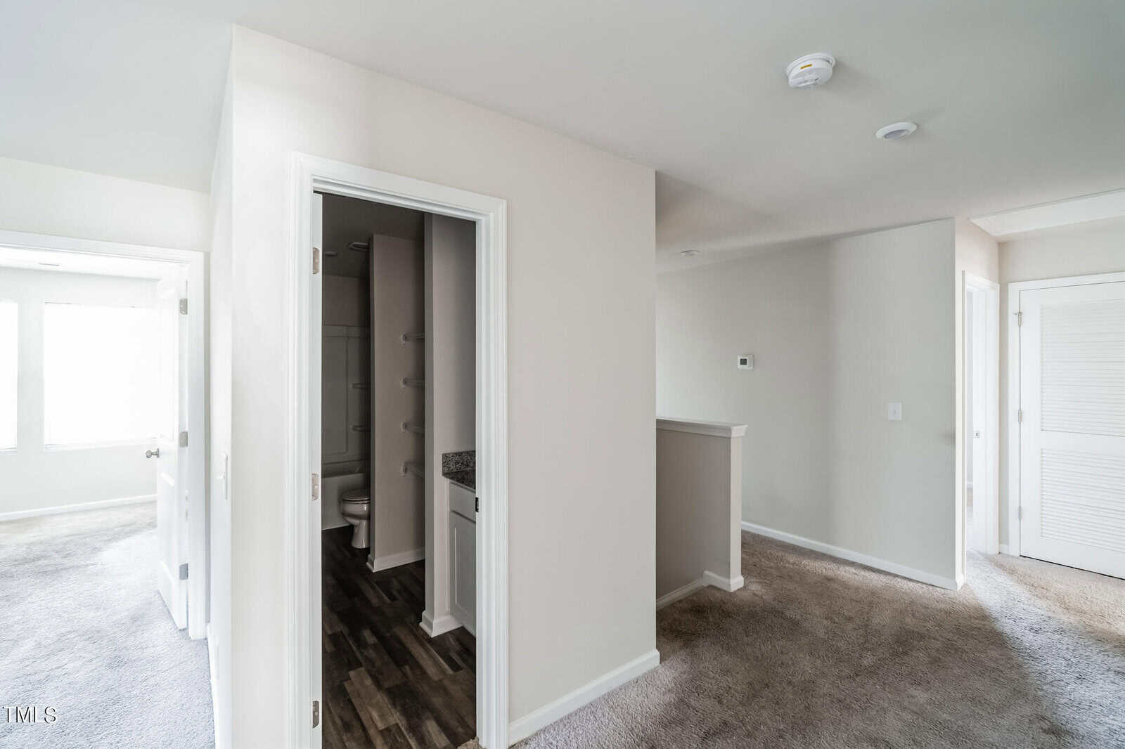 A well-lit hallway in a modern home, featuring beige walls and a mix of hardwood and carpeted floors. The hallway leads to a bathroom on the left with a partially open door, a bright room with a window straight ahead, and a closed door on the right.
