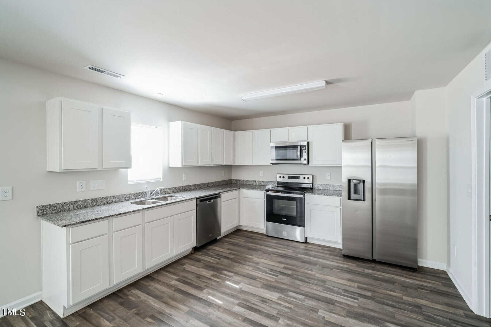 A modern kitchen with white cabinets, a granite countertop, and stainless steel appliances including a refrigerator, oven, microwave, and dishwasher. The room has light-colored walls, gray wood flooring, and a window providing natural light.