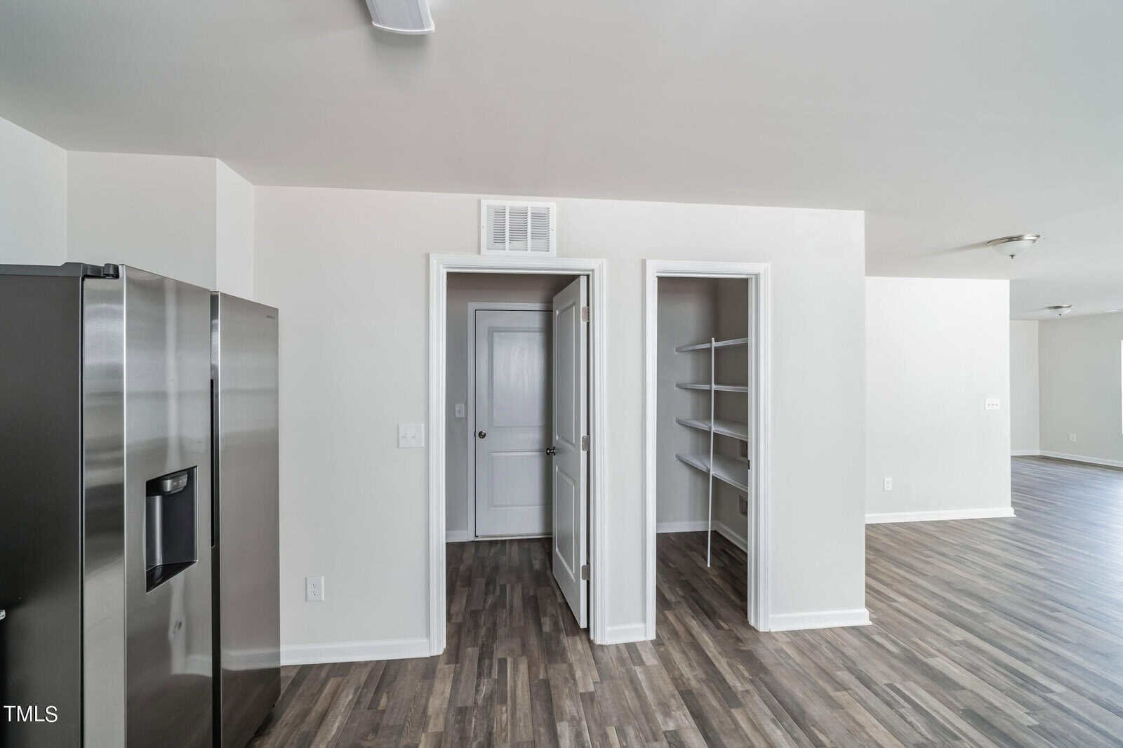 A modern interior showing a kitchen area with a stainless steel refrigerator on the left, white walls, wood flooring, an open pantry with shelves in the center, and a doorway with a closed white door on the right. The background reveals more open living space.