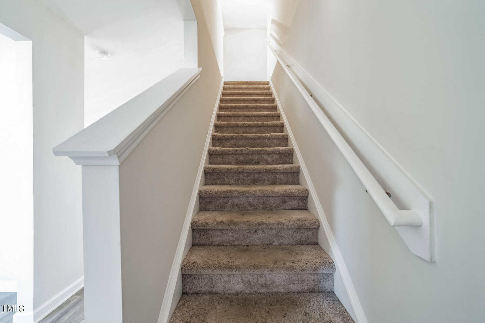 A carpeted staircase with white walls ascends towards a bright light source at the top. A white handrail runs along the right side, with a half-wall on the left. The simple, clean design creates a stark contrast between the carpet and the smooth walls.