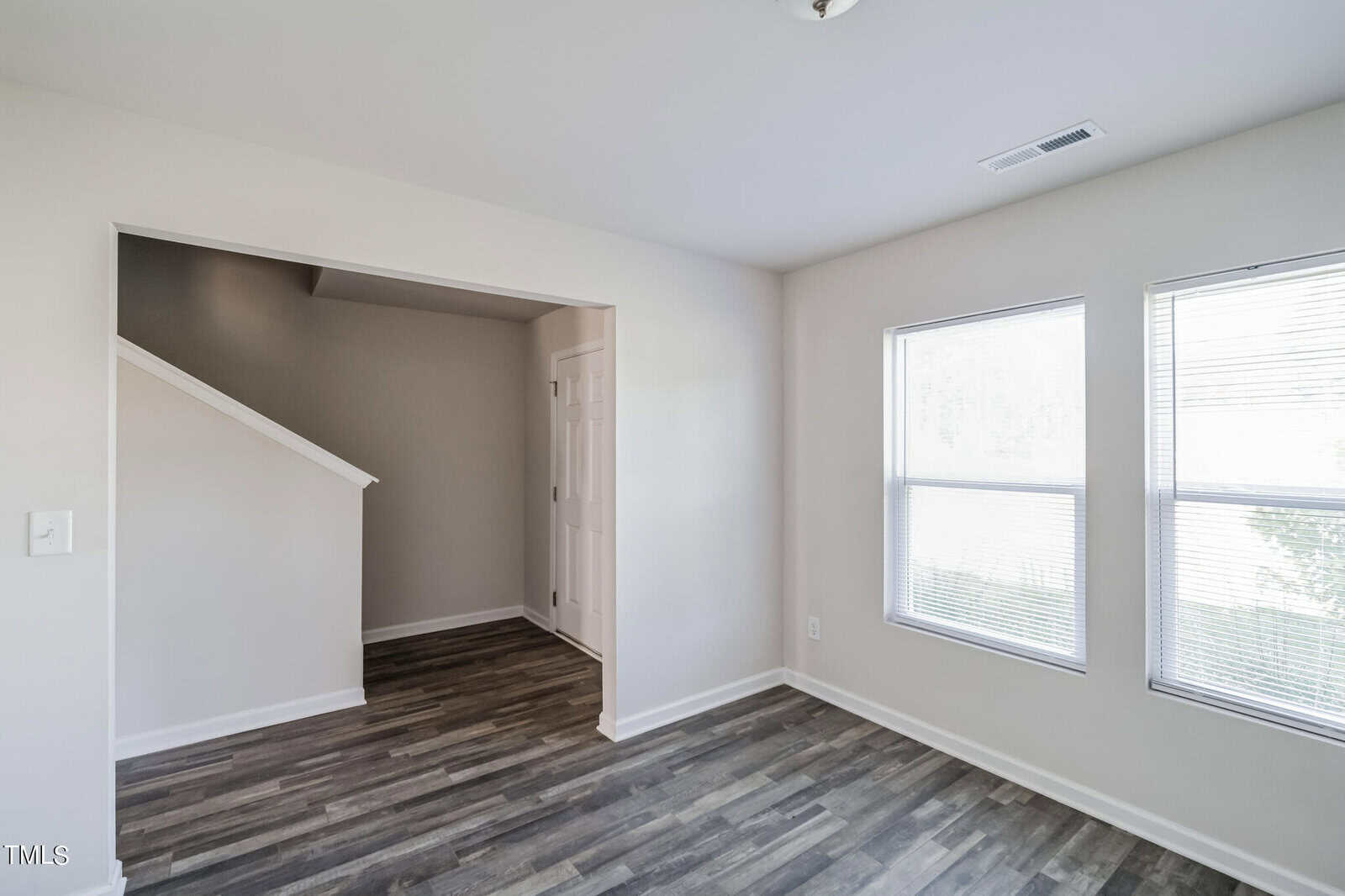 A modern empty room with light gray walls and dark wood flooring. The room features two large windows on the right allowing natural light to enter. In the background, an entryway with stairs is visible, leading to another part of the space.