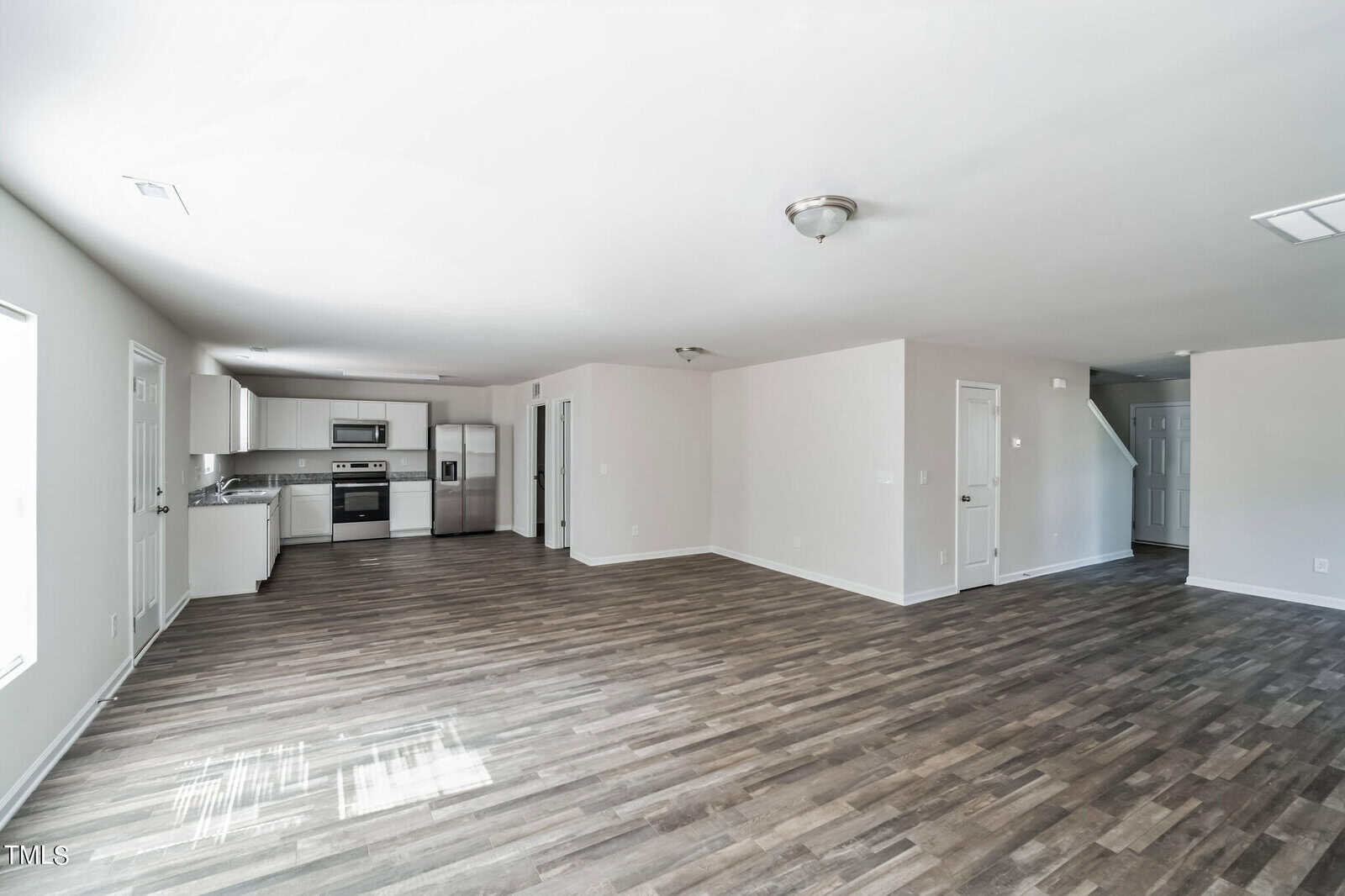 Spacious, modern open-concept living area with light gray walls and dark wood flooring. The room includes a kitchen with stainless steel appliances and white cabinets, a connected dining area, and ample natural light from multiple windows.