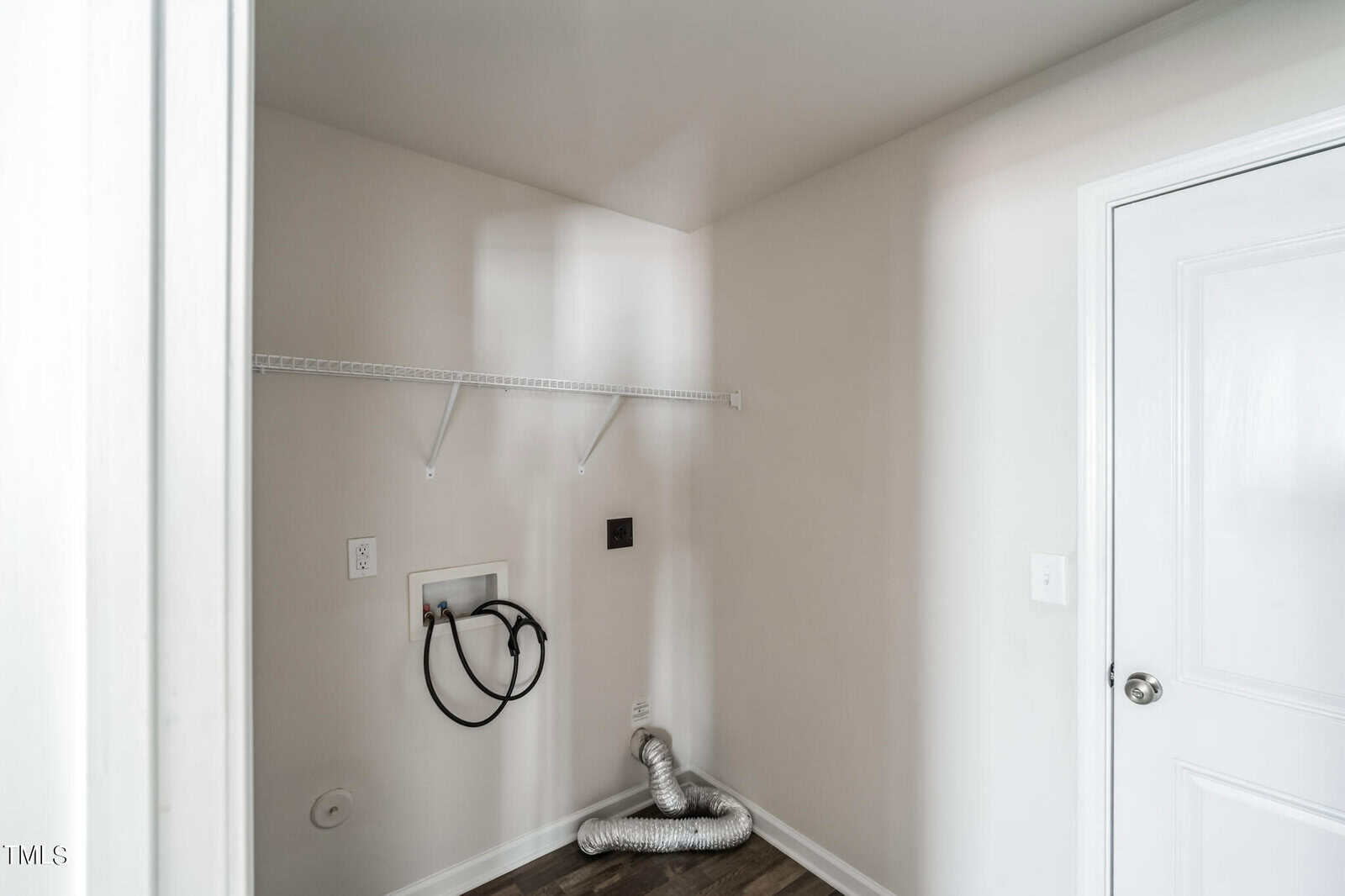 A small laundry room with a single white wire shelf mounted on the wall. Below the shelf, there is plumbing and electrical connections for a washer and dryer. A silver vent hose lies coiled on the laminate floor, and a white door is partially visible on the right.