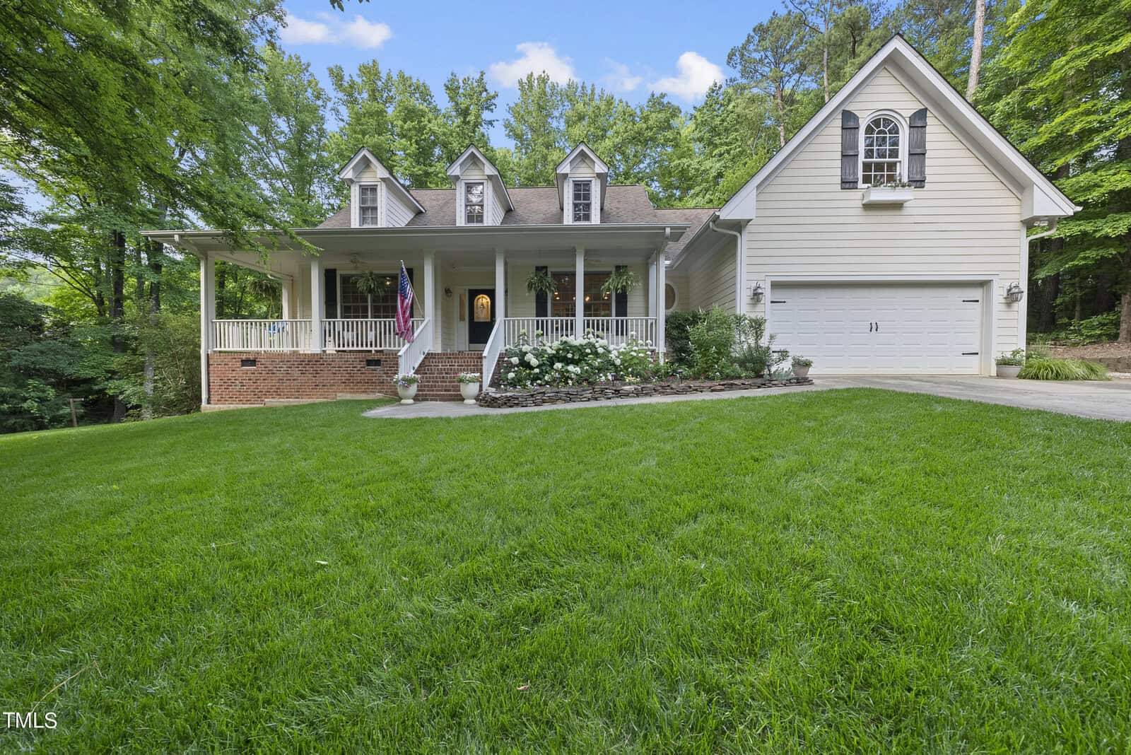 At 568 Timberknoll Lane in Wake Forest, this charming two-story house boasts a white exterior and a front porch with columns adorned by an American flag. The property features a well-manicured lawn, a flower bed, and a driveway leading to a garage, all surrounded by tall green trees under the blue sky.
