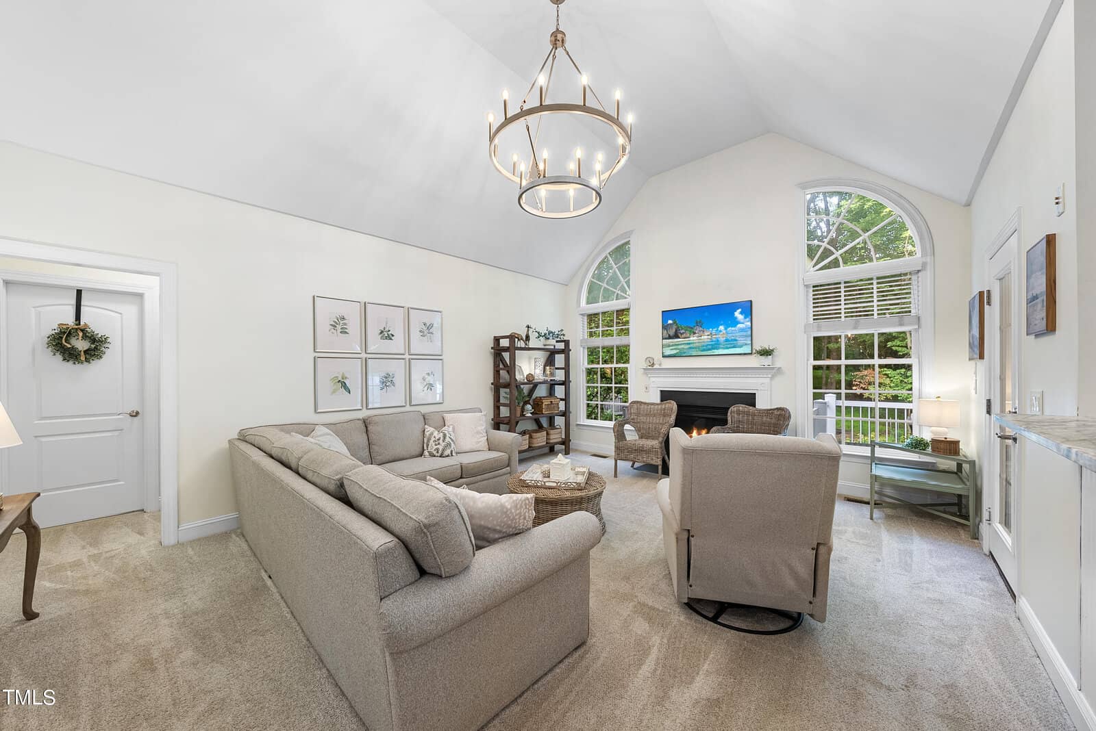 A cozy living room with vaulted ceilings, featuring a large sectional sofa and two armchairs facing a fireplace. A chandelier hangs above, and a flat-screen TV is mounted above the mantel. Large arched windows bring in natural light and greenery views.