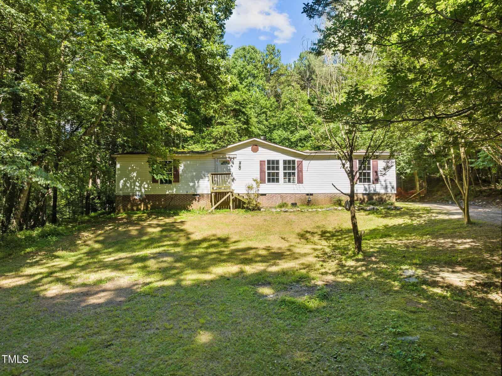 Nestled at 3122 Willow Creek Drive in Wake Forest, this charming single-story house with white siding and a circular window is surrounded by lush trees. A small front porch and steps lead to the entrance, while a dirt path meanders to the right under a sunny, clear sky.
