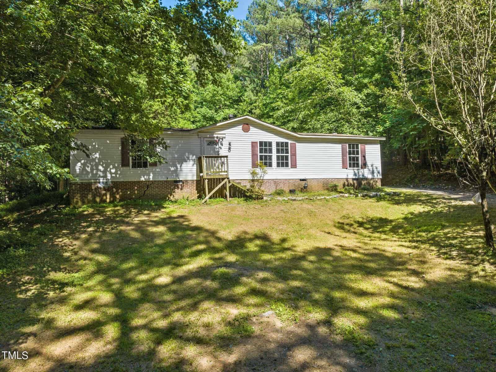A single-story manufactured home with white siding and a small wooden porch is surrounded by grass and trees. The roof is slightly pitched, and there are several windows. The property is in a wooded area, providing shade and greenery.