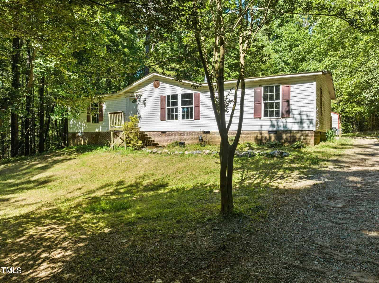 A single-story mobile home with white siding and brown shutters is situated in a wooded area. It has a small front porch and a gravel driveway leading up to the entrance. Surrounding trees provide ample shade and greenery.