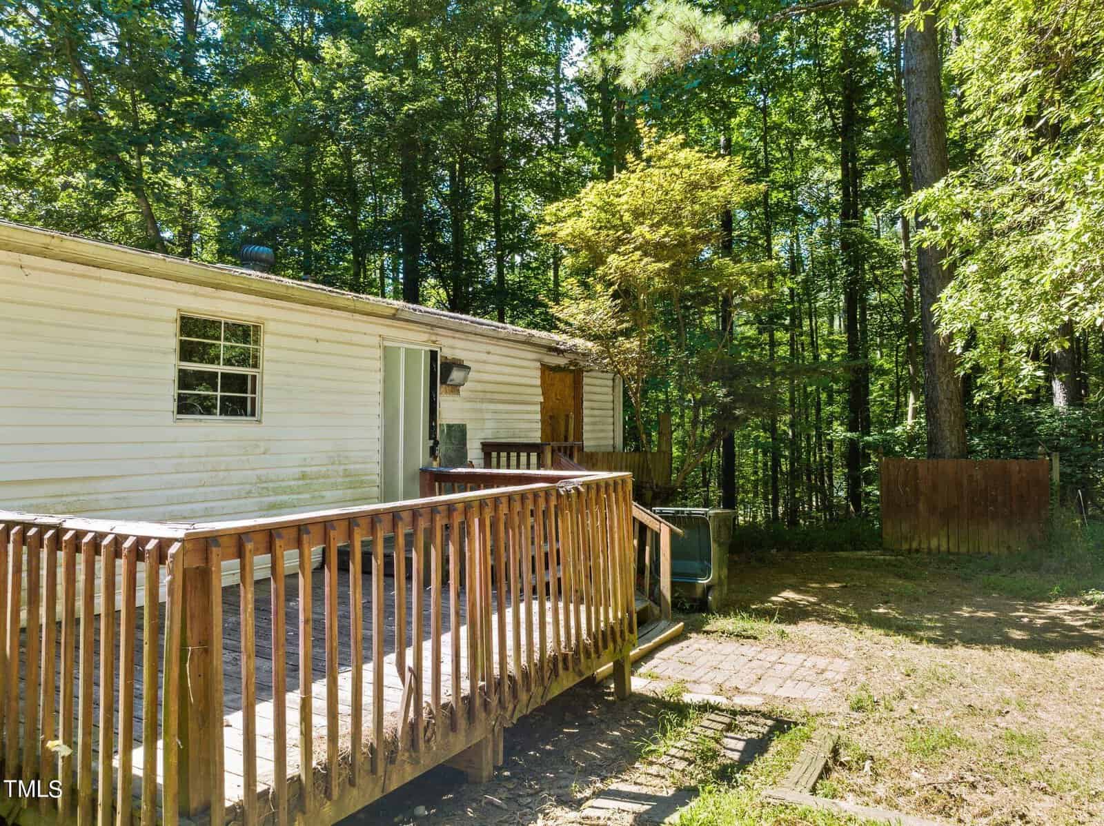 A white mobile home with a wooden ramp entrance is situated in a wooded area. The structure is surrounded by tall green trees and has a small brick patio at its base. A closed window and door are visible on the side of the home.