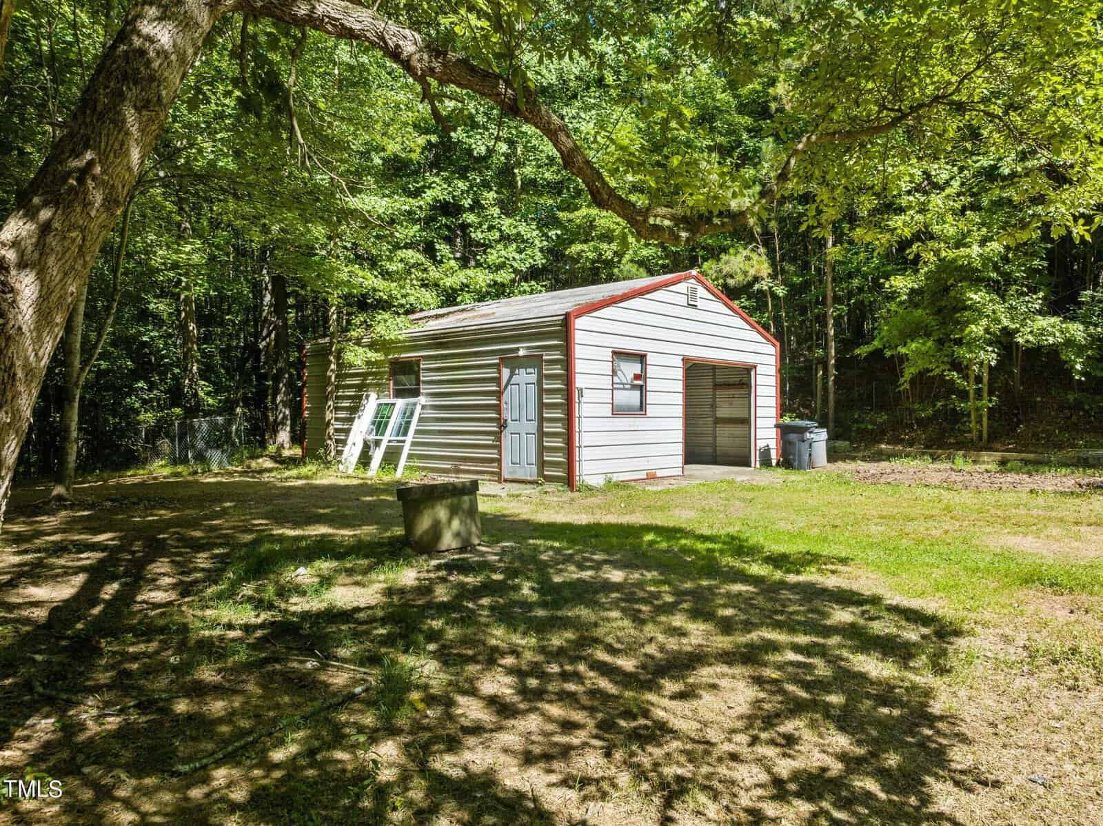 A small, single-story building with light-colored siding and a sloped roof stands surrounded by grass and trees. A tree branch extends overhead, and a few items are placed beside the building. The background is wooded.
