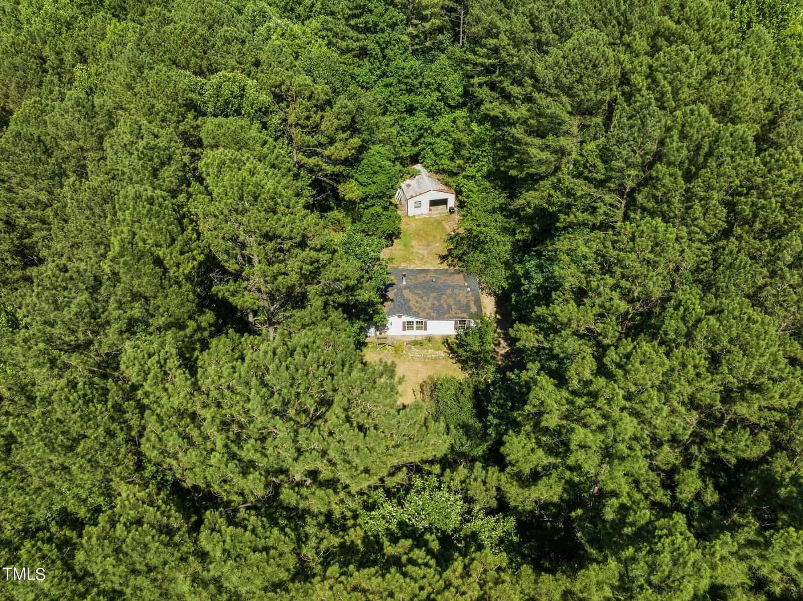 Aerial view of a small white house surrounded by dense green forest. Another smaller structure is visible nearby. The scene is lush and heavily wooded, suggesting a secluded location.