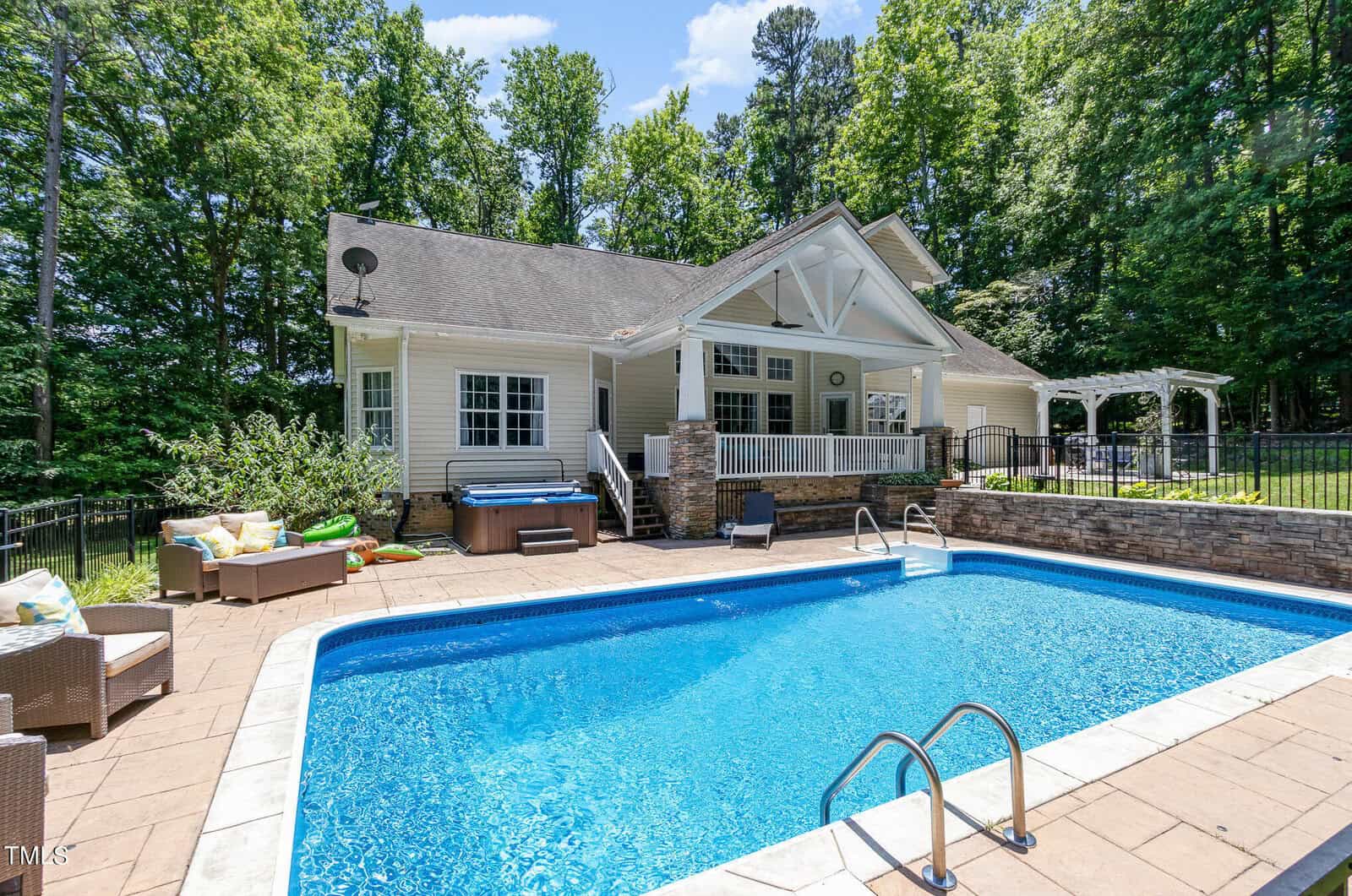 A modern house with a gabled roof sits surrounded by trees on 679 Roberts Chapel Road. In the foreground, there's a blue rectangular pool with a metal ladder. Nearby are a hot tub, outdoor furniture, and a patio. The sky is clear and sunny, creating an idyllic retreat.