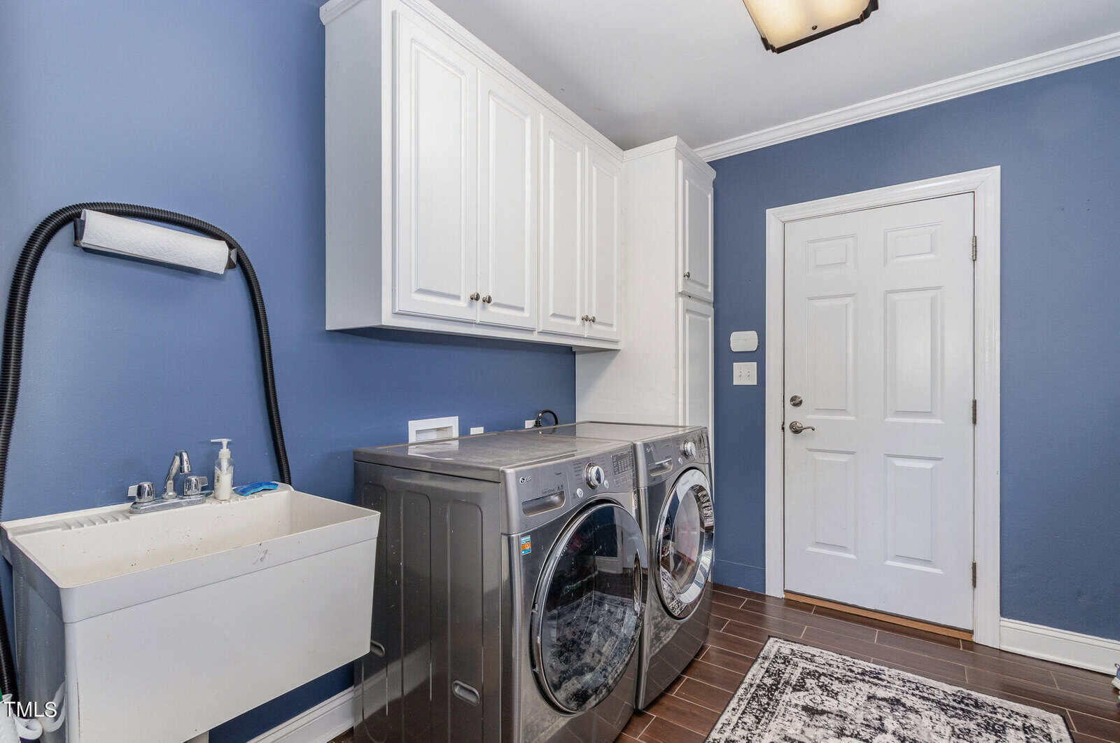 A laundry room with blue walls features a white sink, silver washer and dryer, and white cabinets. A patterned rug lies on dark tiled floors, and a white door is in the background.
