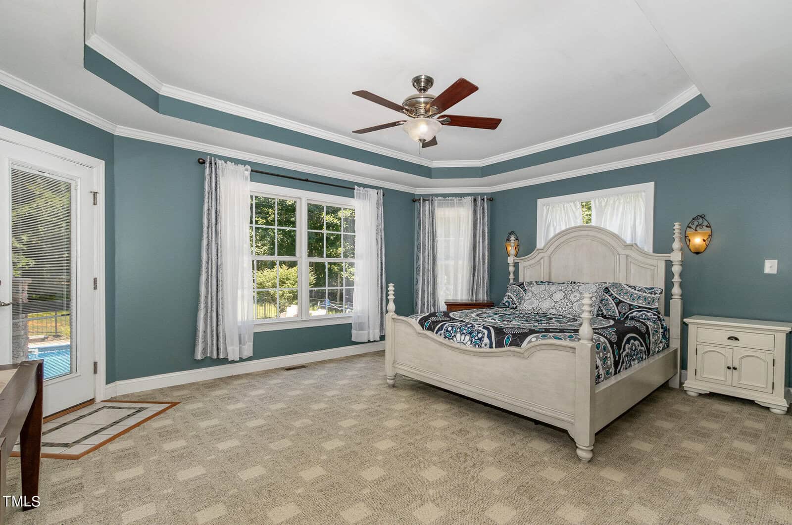 A spacious bedroom with teal walls and a white tray ceiling. A large bed with patterned bedding is placed against a corner. Two windows bring in natural light. A ceiling fan is centered in the room, and a door leads outside. Beige carpet covers the floor.