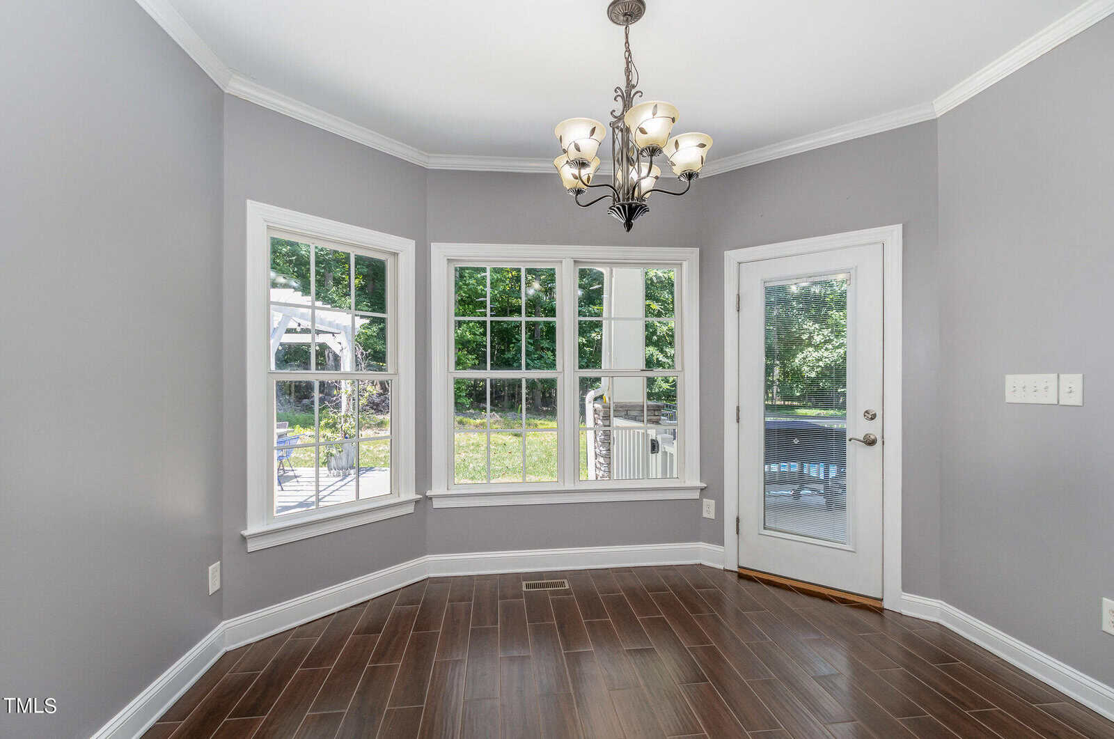 A bright, empty room with gray walls and dark wood flooring. A chandelier hangs from the ceiling. Large windows and a glass door provide a view of a green outdoor space, allowing natural light to fill the room.