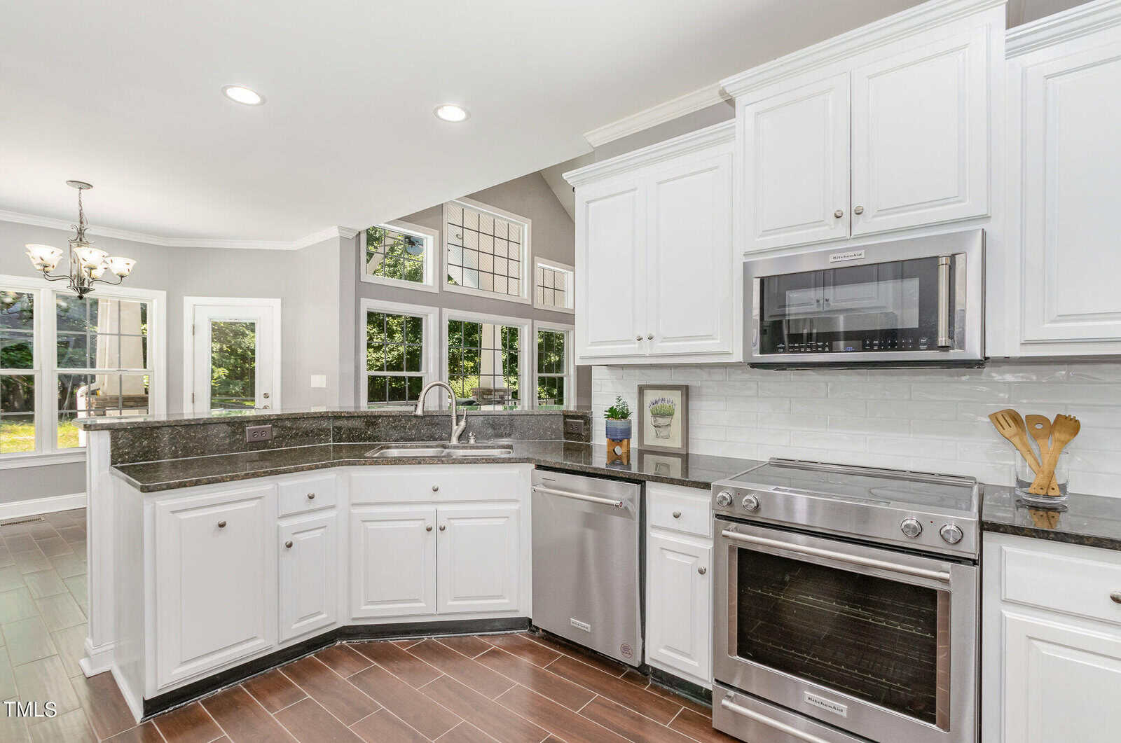 Modern kitchen with white cabinets, black countertops, and stainless steel appliances. Large windows and recessed lighting create a bright atmosphere. There's a view into an adjacent dining area with a chandelier and windows.