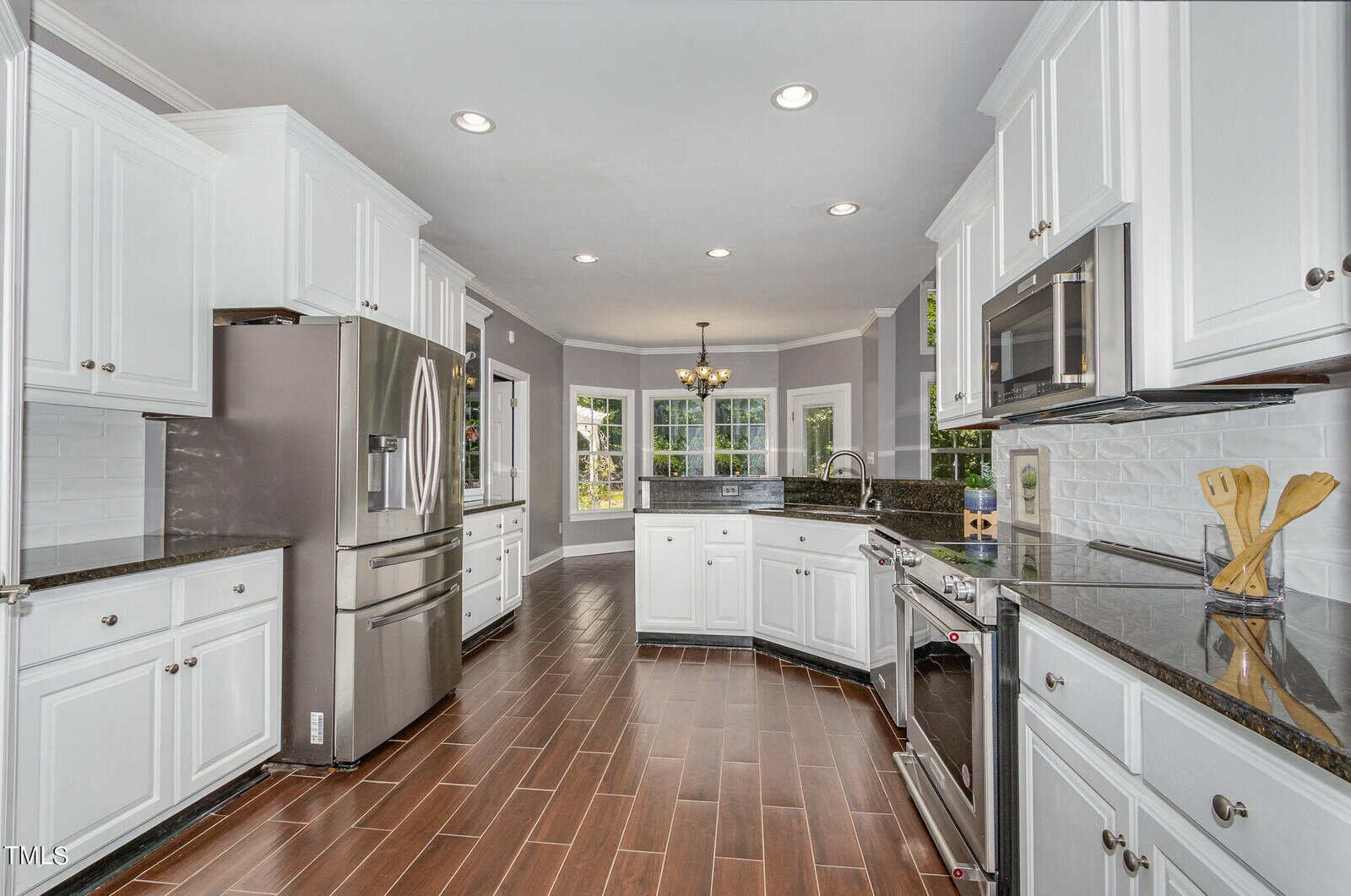 A modern kitchen with white cabinets, stainless steel appliances, and dark granite countertops. The floor is wood-tiled, and a central chandelier hangs by a bay window, providing natural light.