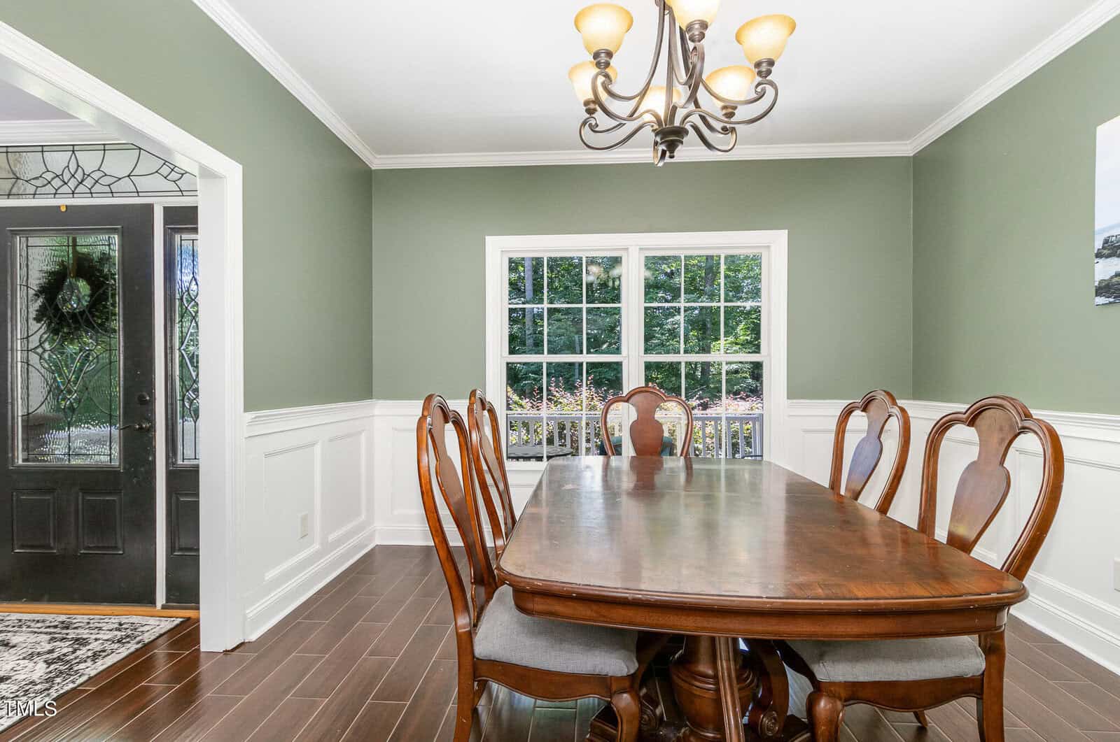 A dining room with green walls, a wooden table, six chairs, and a black chandelier. A large window overlooks greenery outside. A doorway with a wreath on the glass door is visible on the left. Brown wooden flooring complements the decor.