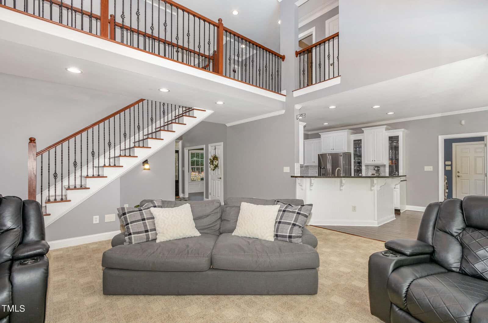 A spacious living room with a gray sofa and armchairs on a beige carpet. The room features an open staircase with wooden railings, a kitchen with white cabinets and a breakfast bar, and high ceilings with recessed lighting.
