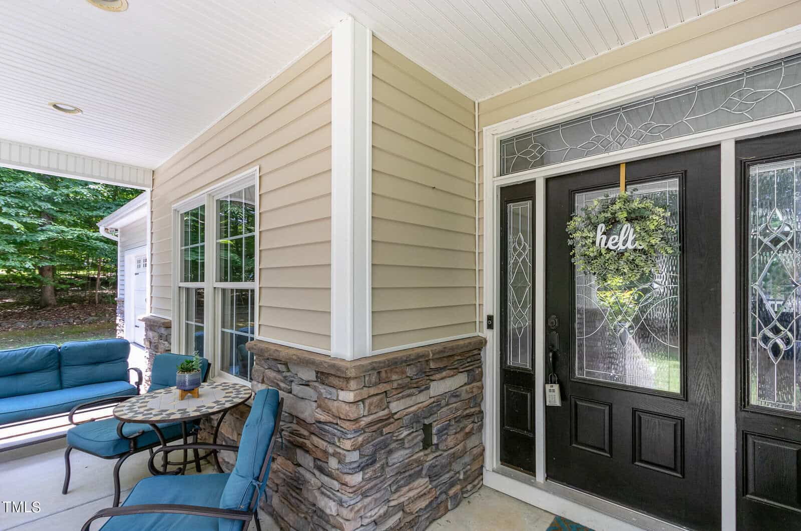 Front porch with a black door, decorative glass panels, and a "hello" wreath. The porch features a stone and siding exterior, a small table with chairs, and a blue cushioned seating area. Trees are visible in the background.