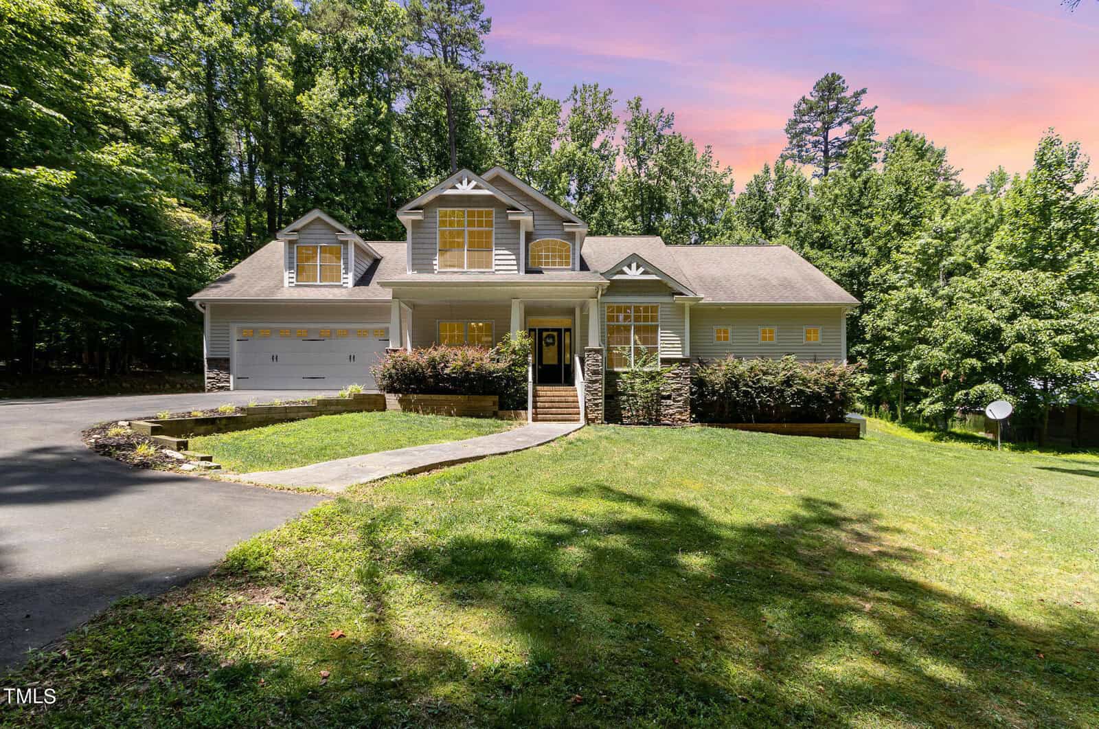 A two-story house with gray siding and white trim is set against a lush, wooded backdrop. The sun sets in the sky, casting a warm glow. The house features symmetrical windows, a double garage, and a curved driveway leading up to the entrance.